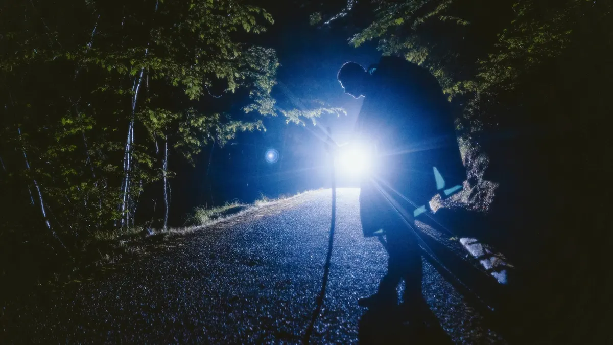 夜の森の道で、人が強い光を放つ懐中電灯を持って立っているシルエット写真。光の筋が前方を照らしている。