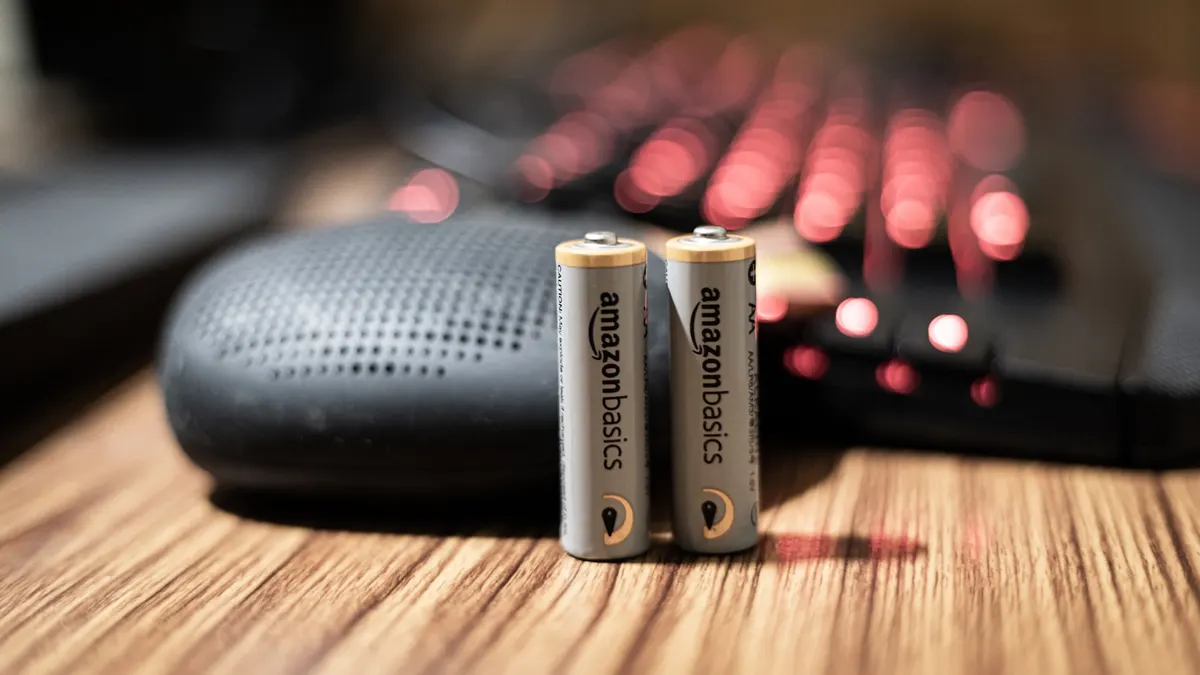 木製のテーブルの上に、Amazonベーシックのアルカリ乾電池が2本立てて置かれている接写。背景はぼやけた赤い光。