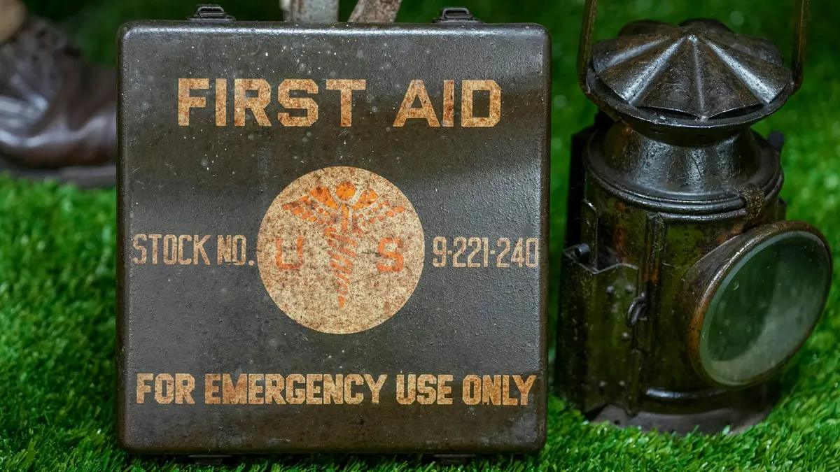 草の上に置かれた、古い金属製のFIRST AIDの箱と、その隣に立つ黒いランタン。