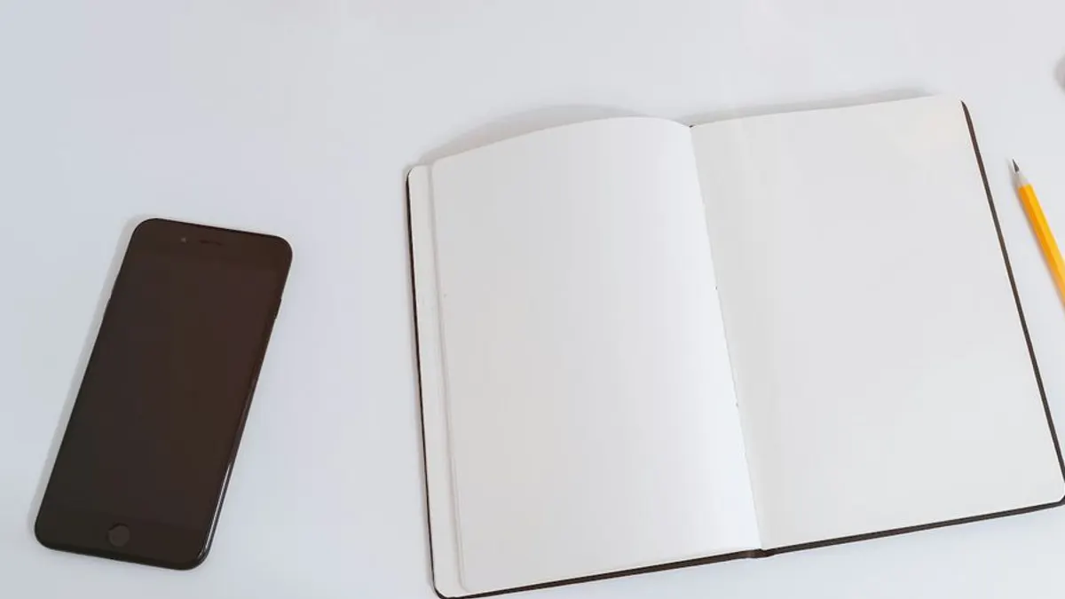 白い机の上に開いた無地の手帳とスマートフォン。固定費の見直し項目を書き出すイメージ。