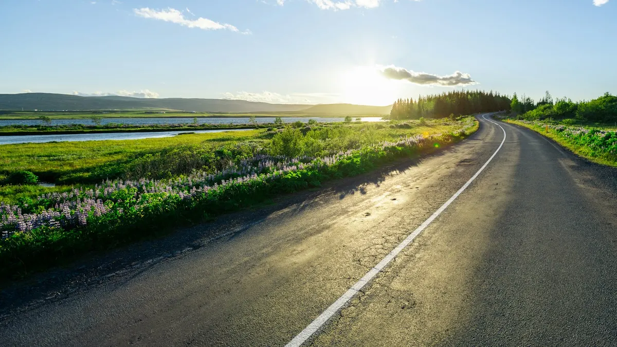 朝日に照らされた田舎道が、緑の野原の間を奥まで伸びている風景写真。10月に向かって進むイメージ。