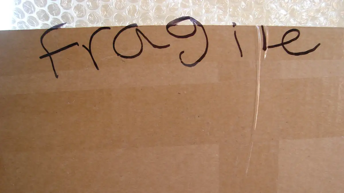 梱包用の段ボールの表面に、黒いマーカーで大きく「fragile」と手書きされている様子。奥には気泡緩衝材が写っている