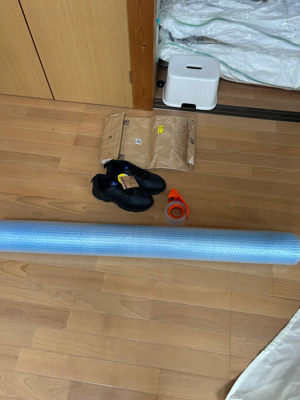 フローリングの床に置かれた黒い安全靴と、過去にAmazonから届いたクラフト紙の配送袋、水色の業務用気泡緩衝材のロール、オレンジ色のテープカッター。