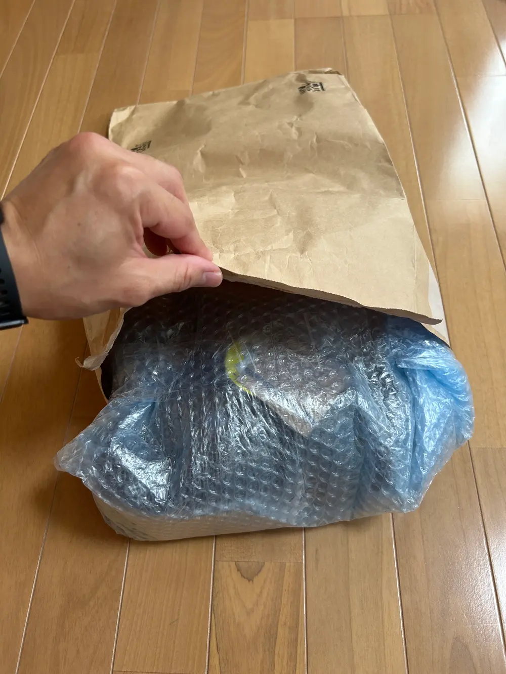 クラフト紙の配送袋の口を手で開き、気泡緩衝材で包んだ安全靴を入れているところ。