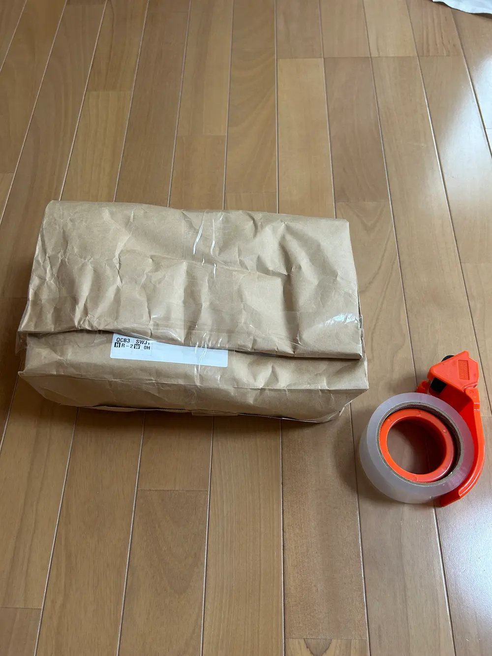 封をした茶色のクラフト紙包みが床に置かれ、右側にオレンジ色のテープカッターに装着された透明の梱包テープが並んでいる。