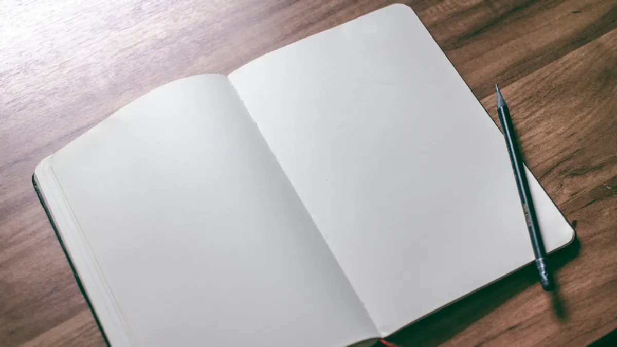 濃い木目の机に開かれた、何も書かれていない白紙のノートと1本の鉛筆。