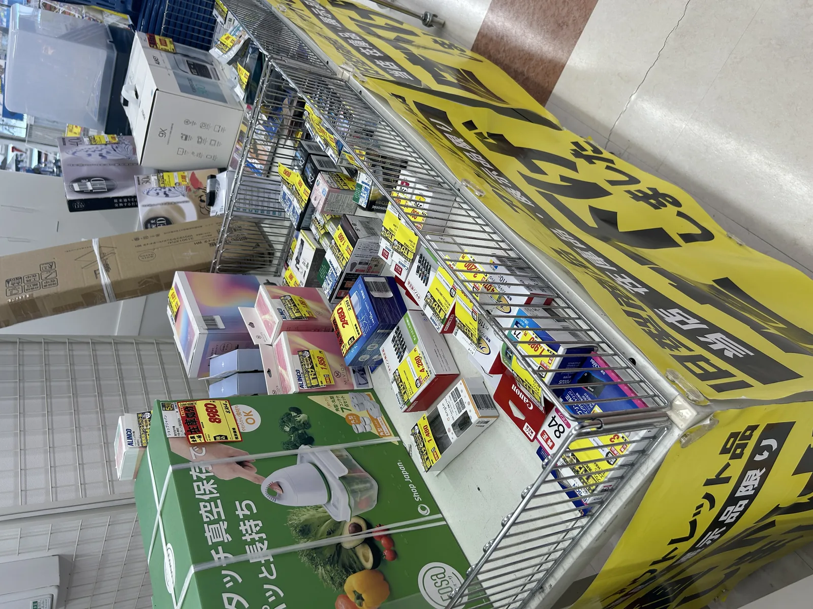 エディオン店内の旧型商品アウトレット棚全体。黄色い「値下げ」バナーが天井から下がり、Panasonic・Canon・EPSON・sonicare・ALINCOなどのメーカー旧モデルが値下げ札とともにワイヤーラックに並ぶ。手前にはShop Japanの緑色パッケージ商品も置かれている