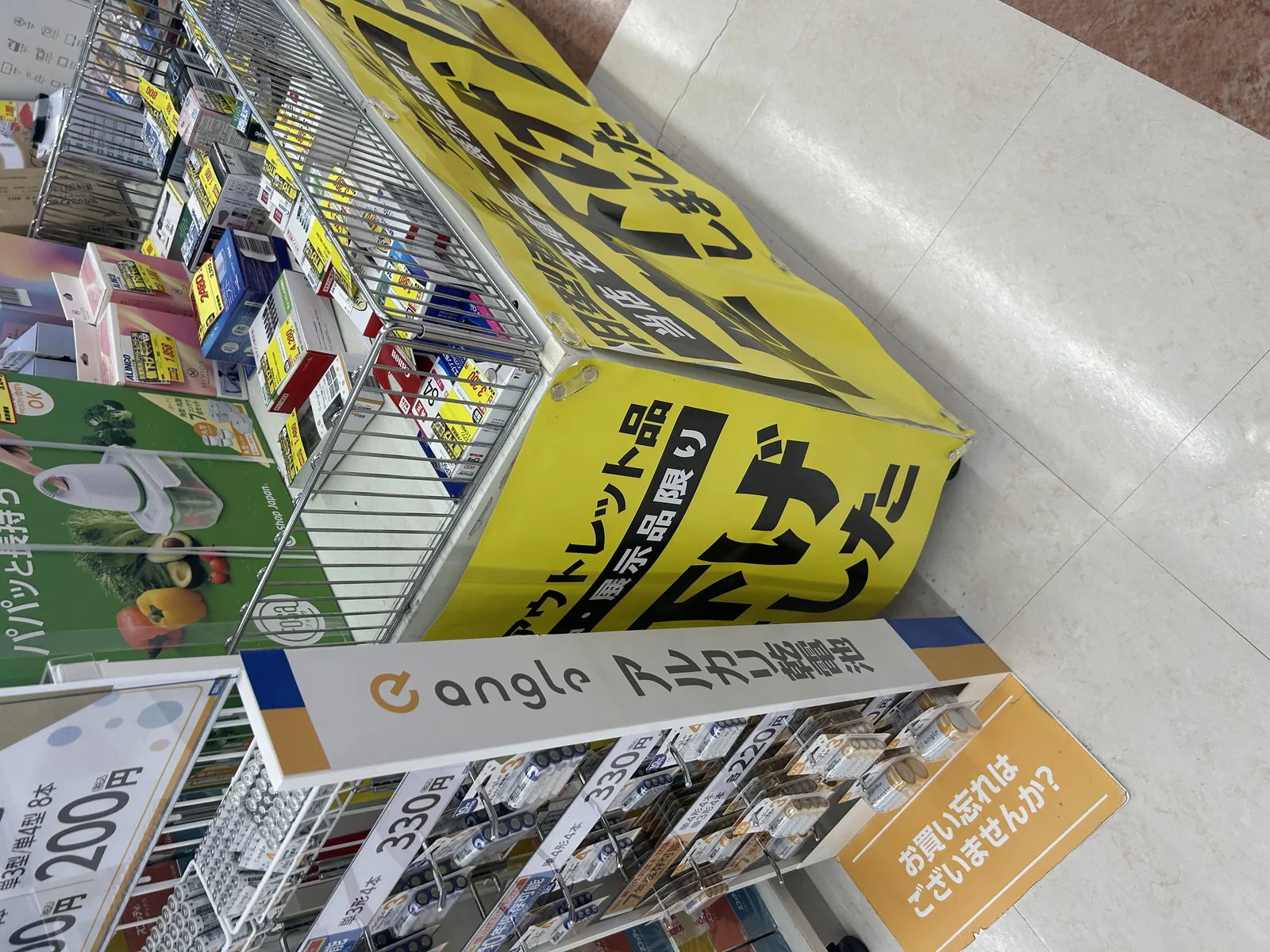 値下げ棚を斜めから撮った拡大写真。黄色バナーの「アウトレット・展示品限り」の文字と、Panasonic・sonicareの旧型番のパッケージ、その隣にエディオンのプライベートブランド『e angle』の乾電池コーナー(単3形 8本200円、単4形 4本330円など)が並ぶ