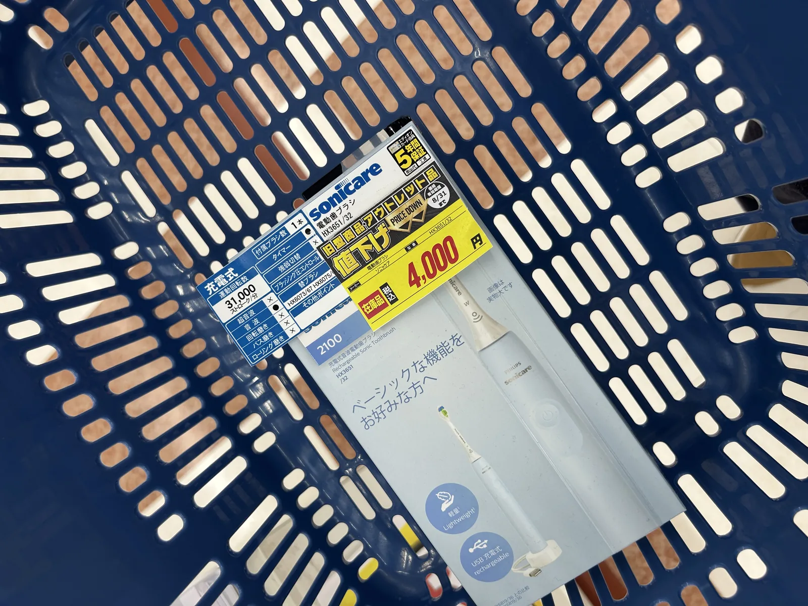 青いエディオンの買い物かごに、フィリップス ソニッケアー2100シリーズ HX3651/32のパッケージが1つ入っている。パッケージには黄色の値下げ札「旧型商品アウトレット PRICE DOWN 8/31まで 電動歯ブラシ ソニッケア 税込¥4,000」と、エディオンカード会員5年保証のシールが貼られている