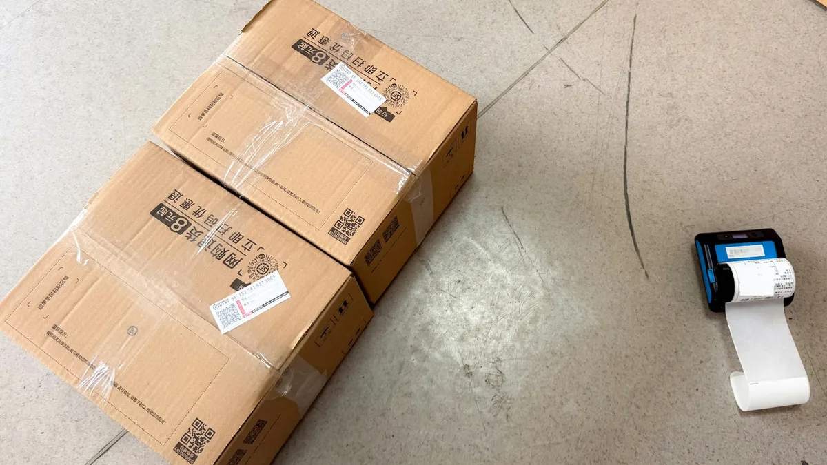 床に置かれた2つの段ボール箱の横に、伝票を印字中の小型ラベルプリンターが置かれている写真。