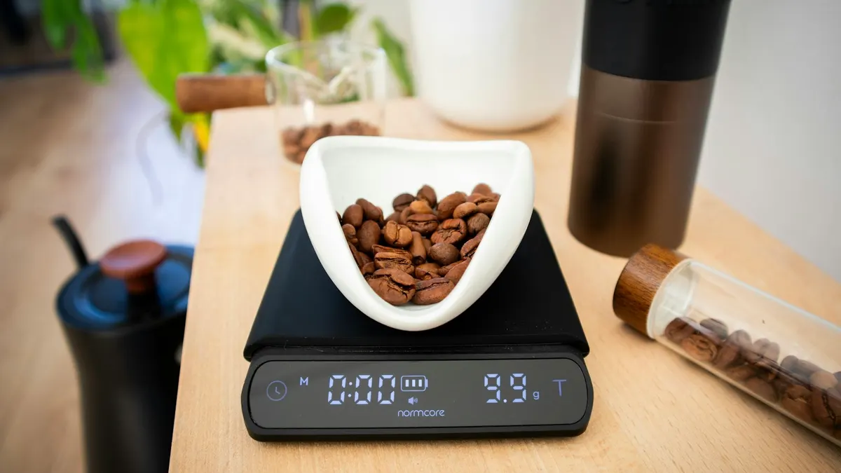 黒いデジタルクッキングスケールの上に、白い器に入ったコーヒー豆が載っていて、表示部に「9.9g」と出ている接写。木製のカウンターの上。