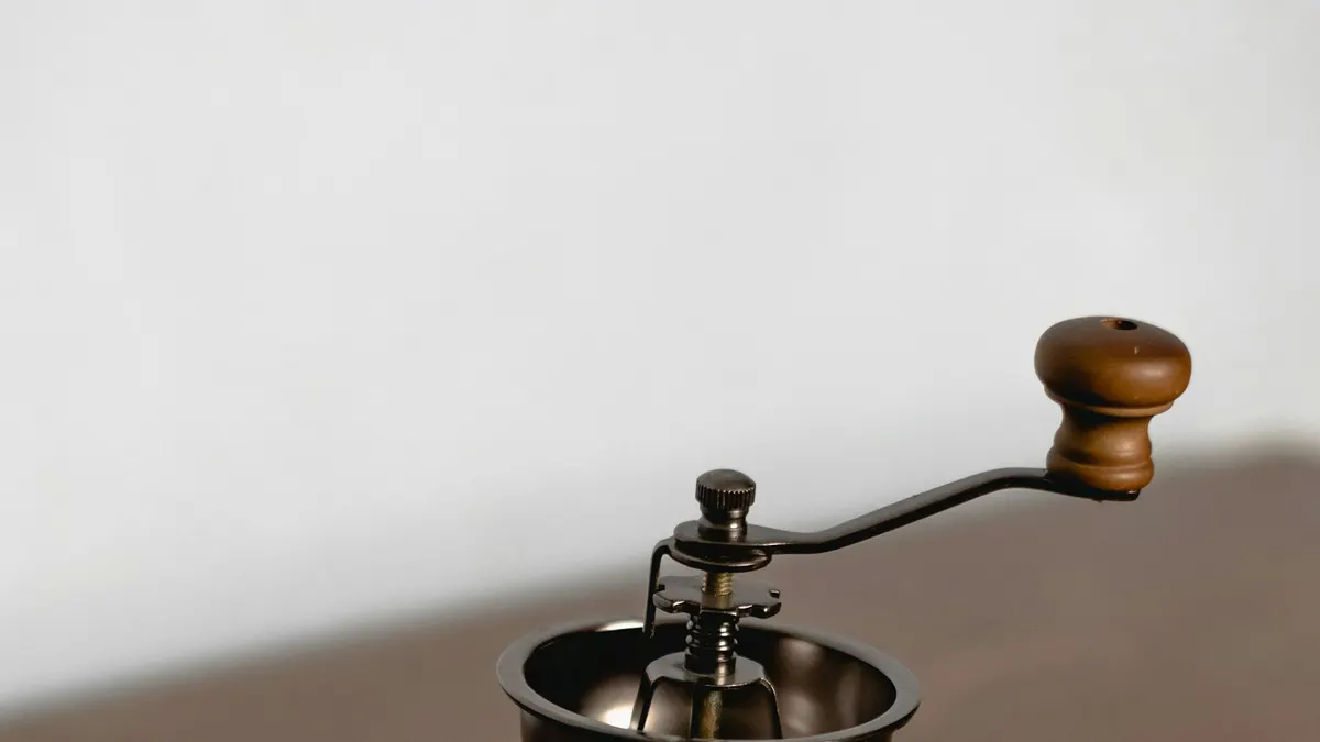 白い壁を背景に、木製のハンドルが付いた小型の手動コーヒーミルが置かれている接写。金属のドーム部分に光が当たっている。