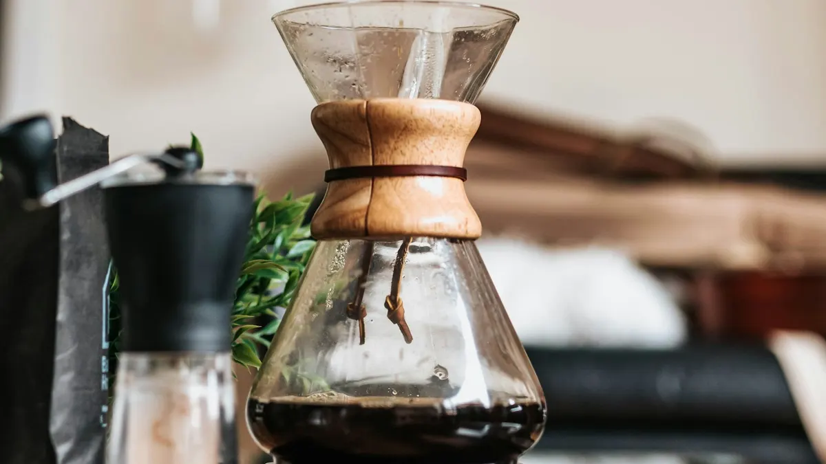 透明なガラスのドリップサーバーにコーヒーが落ちている接写。中央に木製のバンドが巻かれていて、奥にはコーヒーミルと観葉植物がぼやけて写っている。