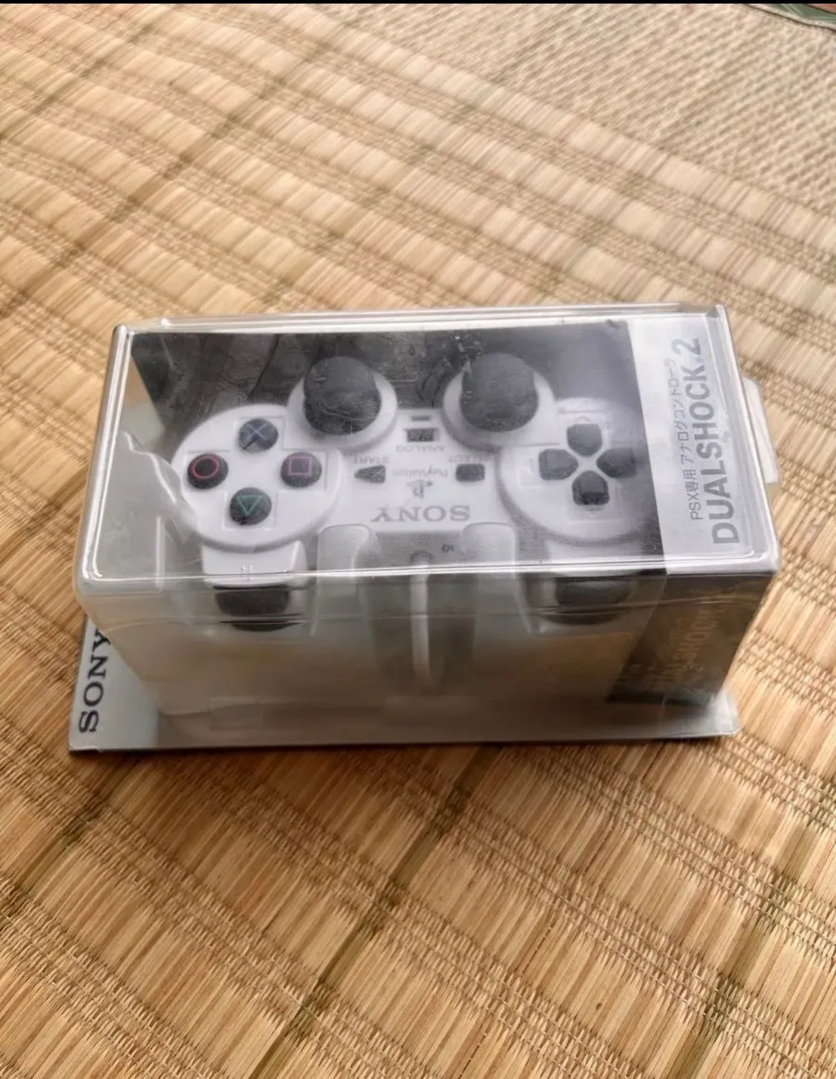 PlayStation純正コントローラ（DUALSHOCK 2・型番DESR-10）の新品未開封品。透明パッケージ越しにホワイトのコントローラ本体が見える表面