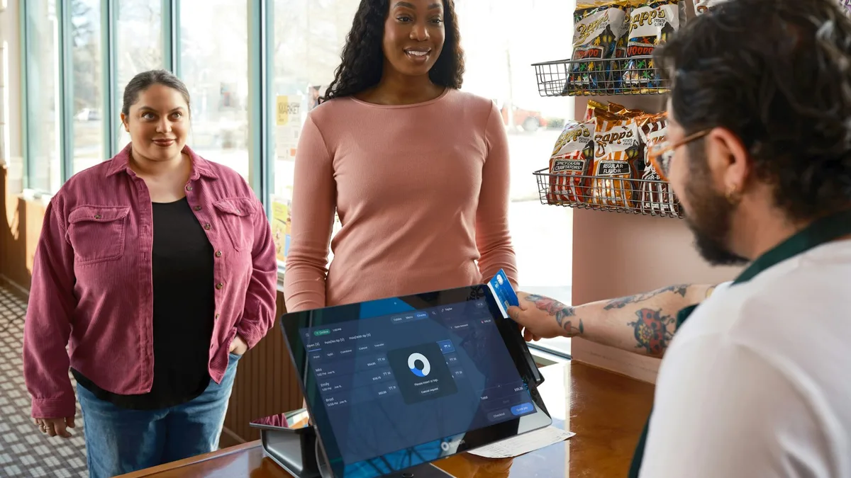 店舗のレジカウンター越しの光景。手前に店員が立ってタブレット型のPOS端末を操作し、カウンターの向こう側に順番を待つ客が並んでいる。右上にスナック菓子の吊り下げラック、左手に大きなガラス窓。