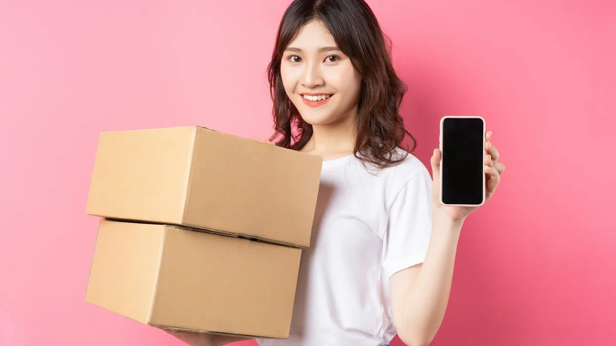ピンク色の背景のスタジオで、白いTシャツを着た若い女性が笑顔で左手にスマートフォンを掲げ、右腕でクラフト色のダンボール箱を2つ抱えている。