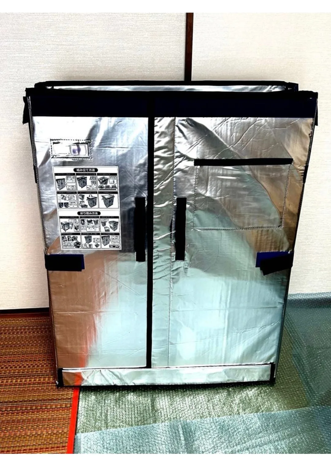 畳の部屋に置かれた、銀色の断熱シート製で紺色の縁取りが入った業務用の折りたたみ式保冷ボックス。天面に黄色い「注意」ラベル、前面にデジタル温度計と組み立て方法・折り畳み方法のイラスト説明シールが貼られ、観音開きの扉と面ファスナーのベルトが付いている。