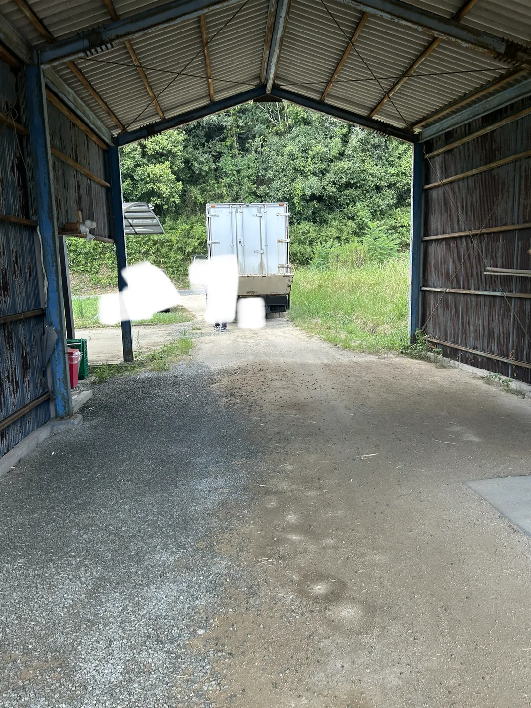 シャッターの開いた倉庫のような建屋の内部から外を見た風景。奥に白いトラックの荷台後部が停まっており、砂利敷きの通路と周囲の木々が写っている。トラックの手前に立つ人物2名はプライバシー保護のため白く加工されている。