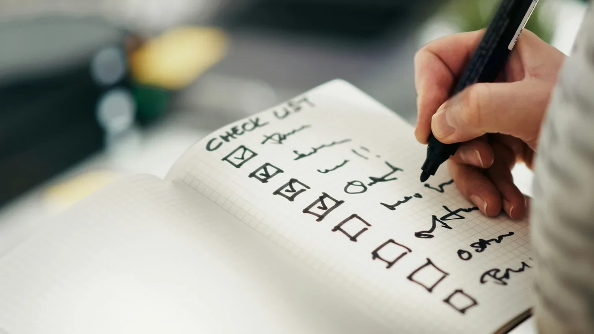 「CHECK LIST」と書かれたノートにチェックを入れる手元。中間チェック完了・継続のイメージ。