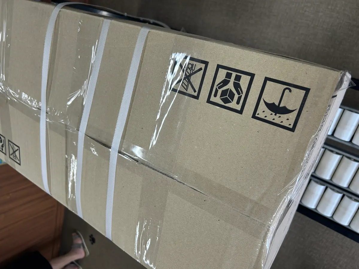 ガムテープで封をした大きな段ボール箱。上面に取扱注意のマークが並び、オレンジ色のテープカッターが載っている。
