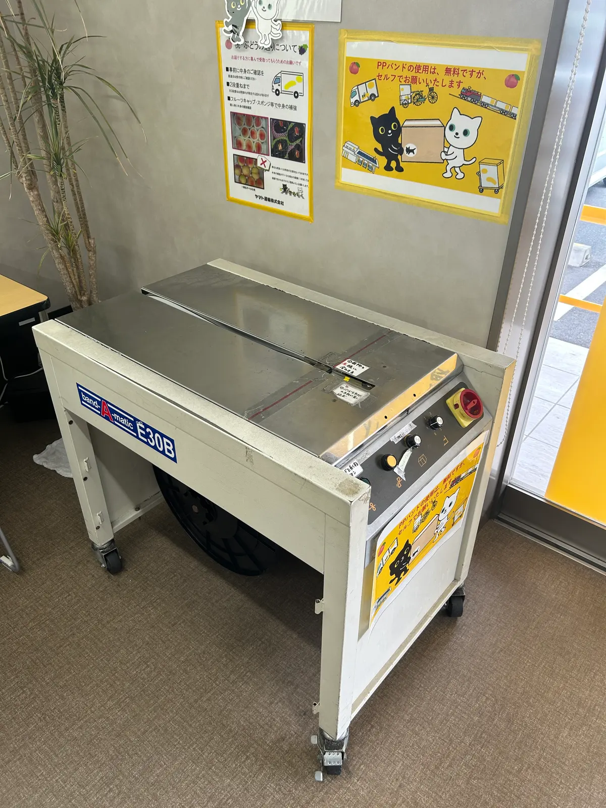 ヤマト運輸の営業所内に置かれたステンレス製のPPバンド結束機（band-A-matic E30B）。壁の黄色い貼り紙に猫のイラストとともに「PPバンドの使用は、無料ですが、セルフでお願いいたします」と書かれている。