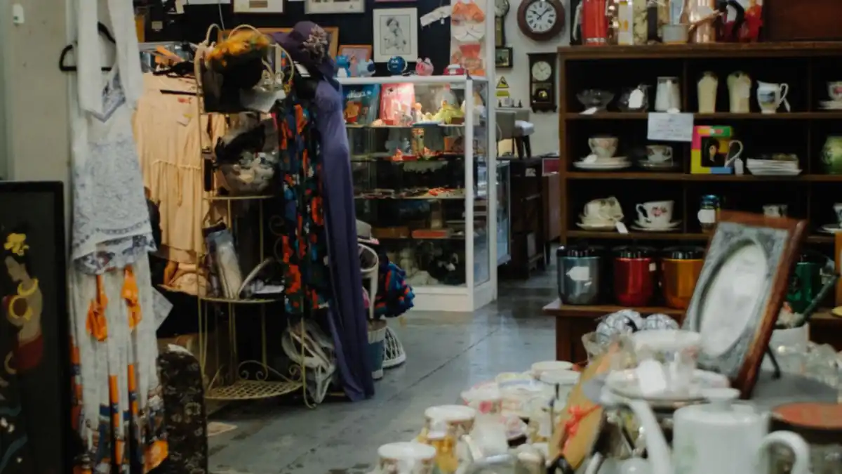 中古品を扱うリサイクルショップの店内を、通路の奥に向かって撮った写真。左手には古着がハンガーラックに掛かり、中央にガラスの陳列ケース、右手の木製の棚には中古の食器やカップ、額縁、缶がぎっしり並んでいる。手前のテーブルにも白い食器類が山積みになっている。人物は写っていない。