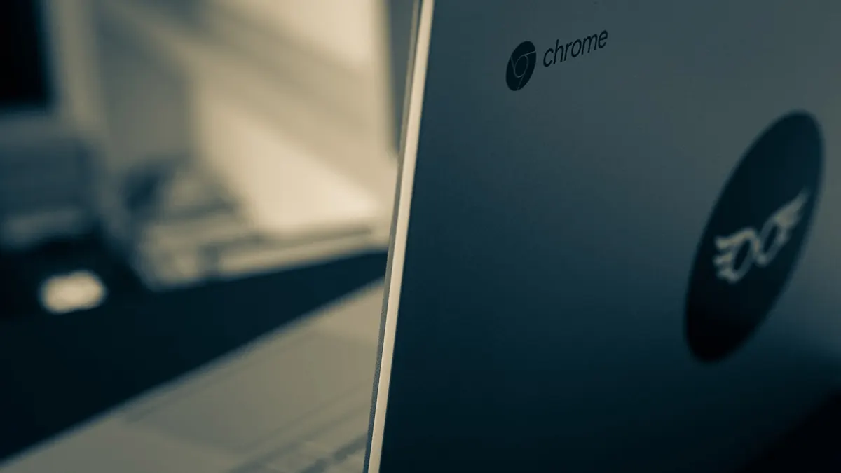 ノートパソコンの天板に入った「chrome」のロゴを斜めから接写した写真。青緑がかった暗いトーンで、奥のデスクはぼやけている。