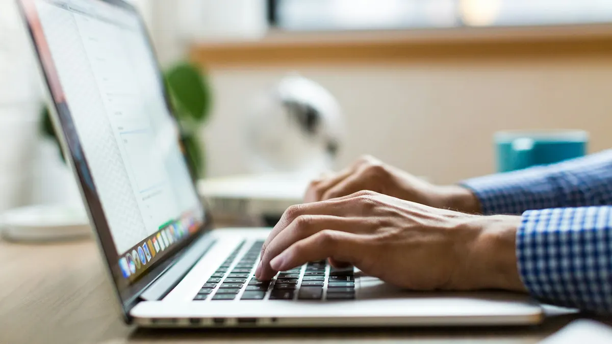 明るい窓辺の木のデスクで、青いチェック柄のシャツを着た人がノートパソコンのキーボードを打っている手元。奥に観葉植物とデスクライトがぼやけて見える。