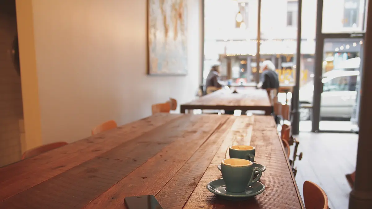 木の長テーブルに置かれた2つのコーヒーカップ。奥の窓から街の光が入るカフェの店内