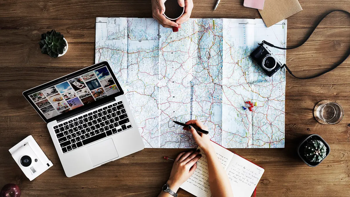 木のテーブルに地図を広げ、ペンで印をつけながらノートに書き込む手元。ノートパソコンとコーヒーが並んでいる