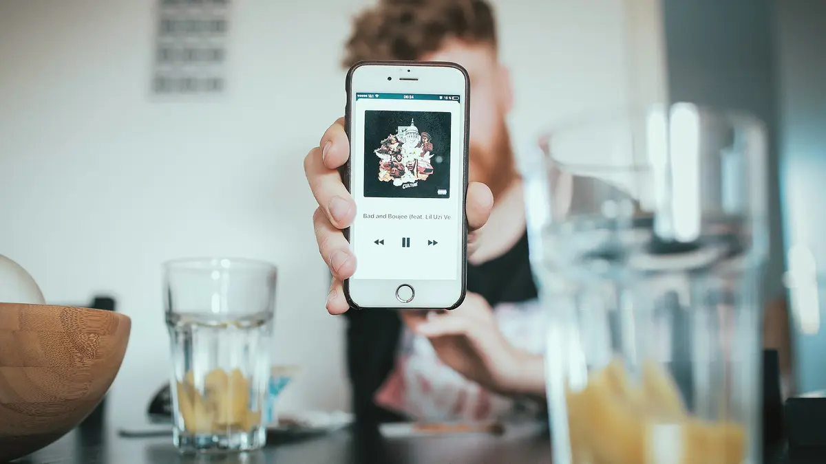 手に持ったスマートフォンの画面をカメラに向けて掲げている人物。背景に人物の顔がぼやけて写っている