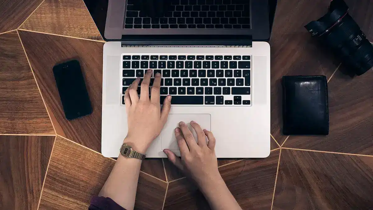 木目の机の上でノートパソコンのキーボードに手を置いている人物。スマートフォンと財布が並んでいる