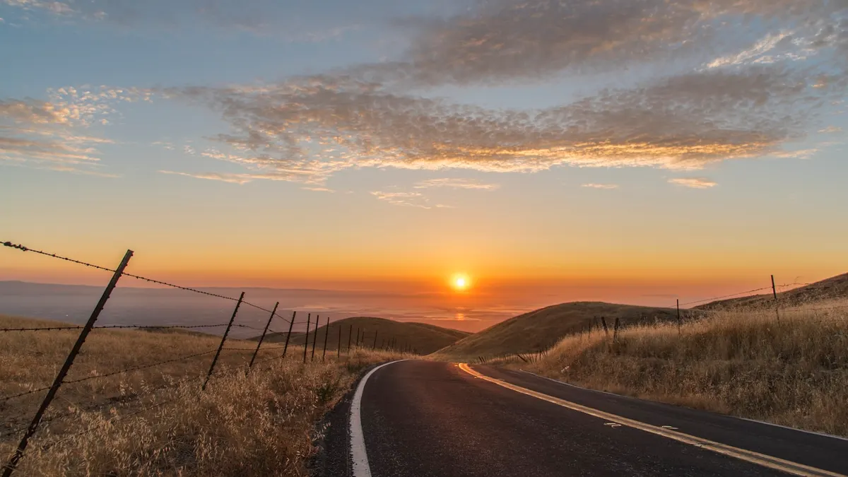 夜明けの空の下、乾いた草地の丘を貫いて延びる2車線の舗装道路。地平線近くにオレンジ色の太陽が昇り、道路の左側に有刺鉄線の柵が続いている。