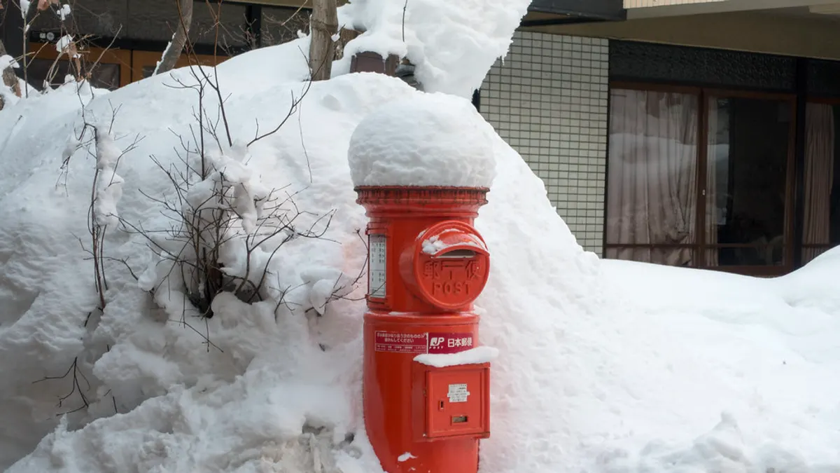 雪が積もった街角に立つ赤い日本郵便のポスト。天面に雪が帽子のように積もり、投函口には「郵便 POST」の文字が見える。背景に格子模様のタイル壁と窓がある。