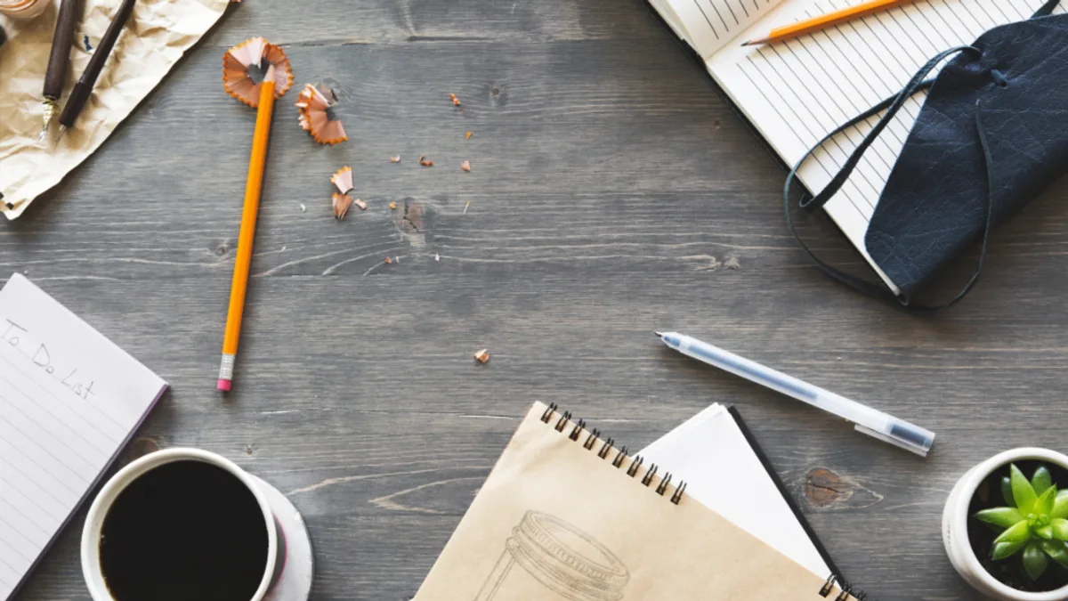 グレーがかった木目のデスクを真上から見た俯瞰。黒いコーヒーの入ったカップ、「To Do List」と書かれた白いメモ帳、罫線ノート、オレンジ色の鉛筆と削りかす、多肉植物の小さな鉢が並んでいる。