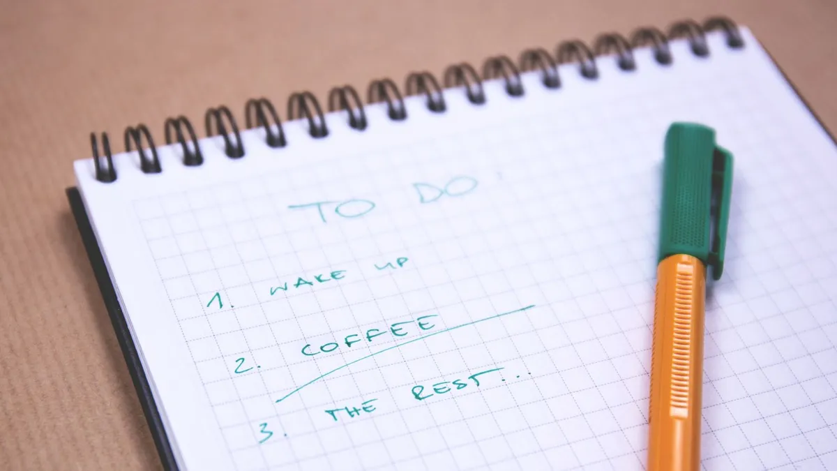 クラフト紙色の机の上で、方眼のリングメモに緑色のペンで「TO DO」と手書きされ、オレンジ色のボールペンが添えられたクローズアップ。