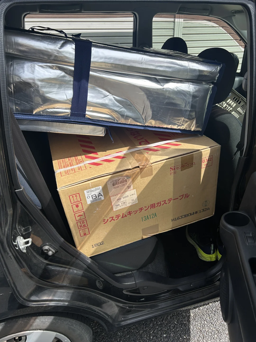 車の後部座席に積み上げた仕入れ品。システムキッチン用ガステーブルの段ボール箱の上に、銀色の保冷バッグが乗っている