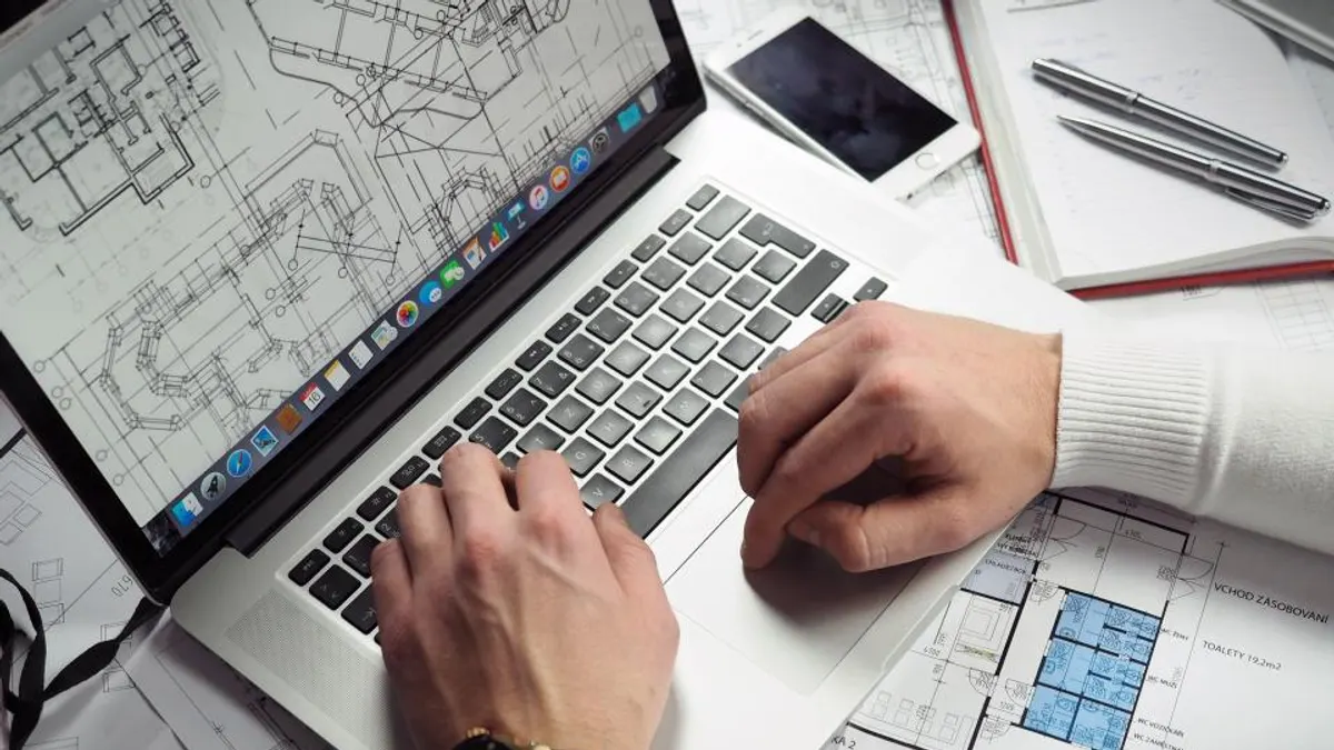ノートPCのキーボードに両手を置いて作業している手元。机の上には図面のような書類、スマートフォン、ペン、ノートが広げられている