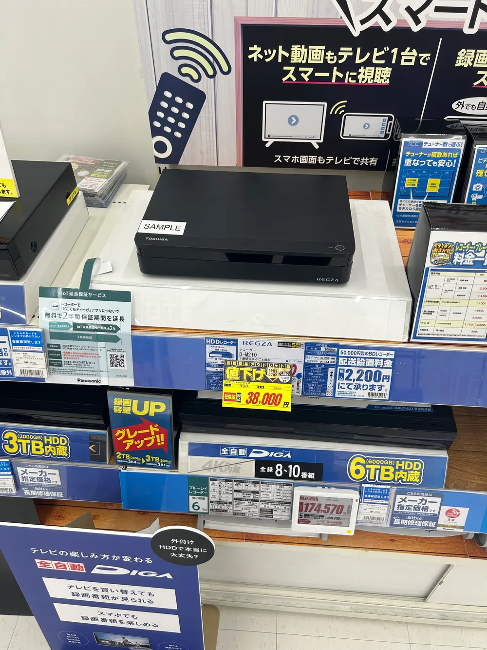 家電量販店の棚に展示されたTOSHIBA REGZAのHDDレコーダー本体。天面に白いSAMPLEシールが貼られ、すぐ下の棚に「旧型商品アウトレット品 値下げ 在庫品 税込38,000円」の黄色い値札が下がっている。