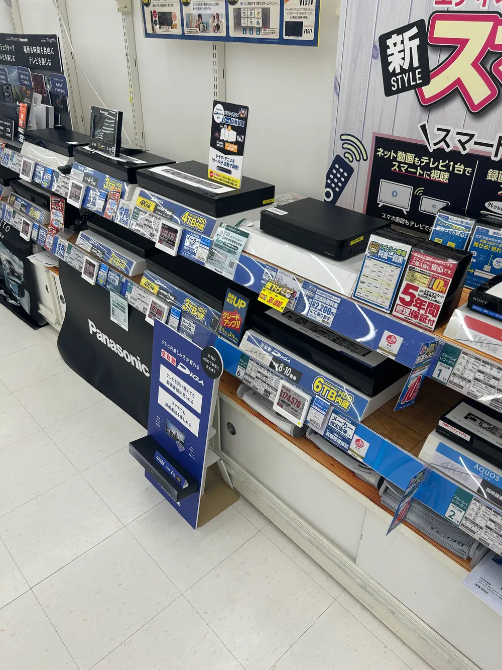 レコーダー売り場のワイドショット。Panasonic DIGAなど他社製品が青と白の棚札とともに整然と並ぶ中で、REGZAの棚にだけ黄色い38,000円の値下げ札が差し込まれている。