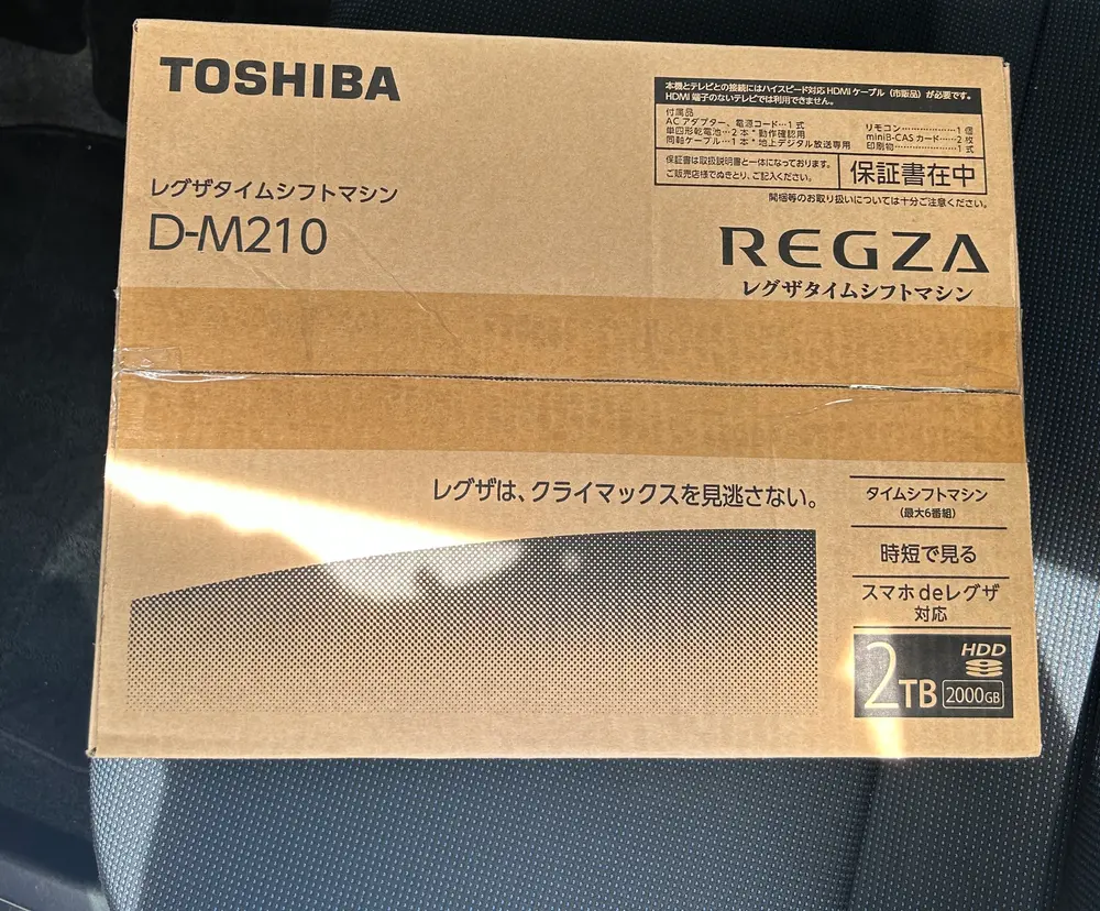 車のシートに置かれたTOSHIBA REGZA タイムシフトマシン D-M210の外箱。段ボールの合わせ目のテープに開封跡があり、右上に付属品リストと「保証書在中」の表示、右下に「2TB 2000GB HDD」の記載がある。