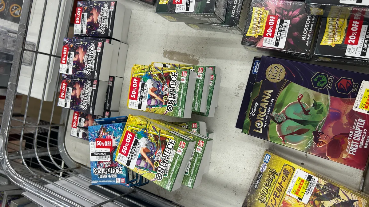 店舗のトレーディングカード売場の棚を斜め上から撮影した写真。手前の段に赤い「50%OFF」ラベルと550円(税込)の値札が付いたONE PIECEカードゲームのスタートデッキが色違いで数種類、複数個ずつ積まれている。奥の段には「20%OFF」ラベルの付いたカードスリーブの箱や、別のトレーディングカードゲームの商品が並んでいる。