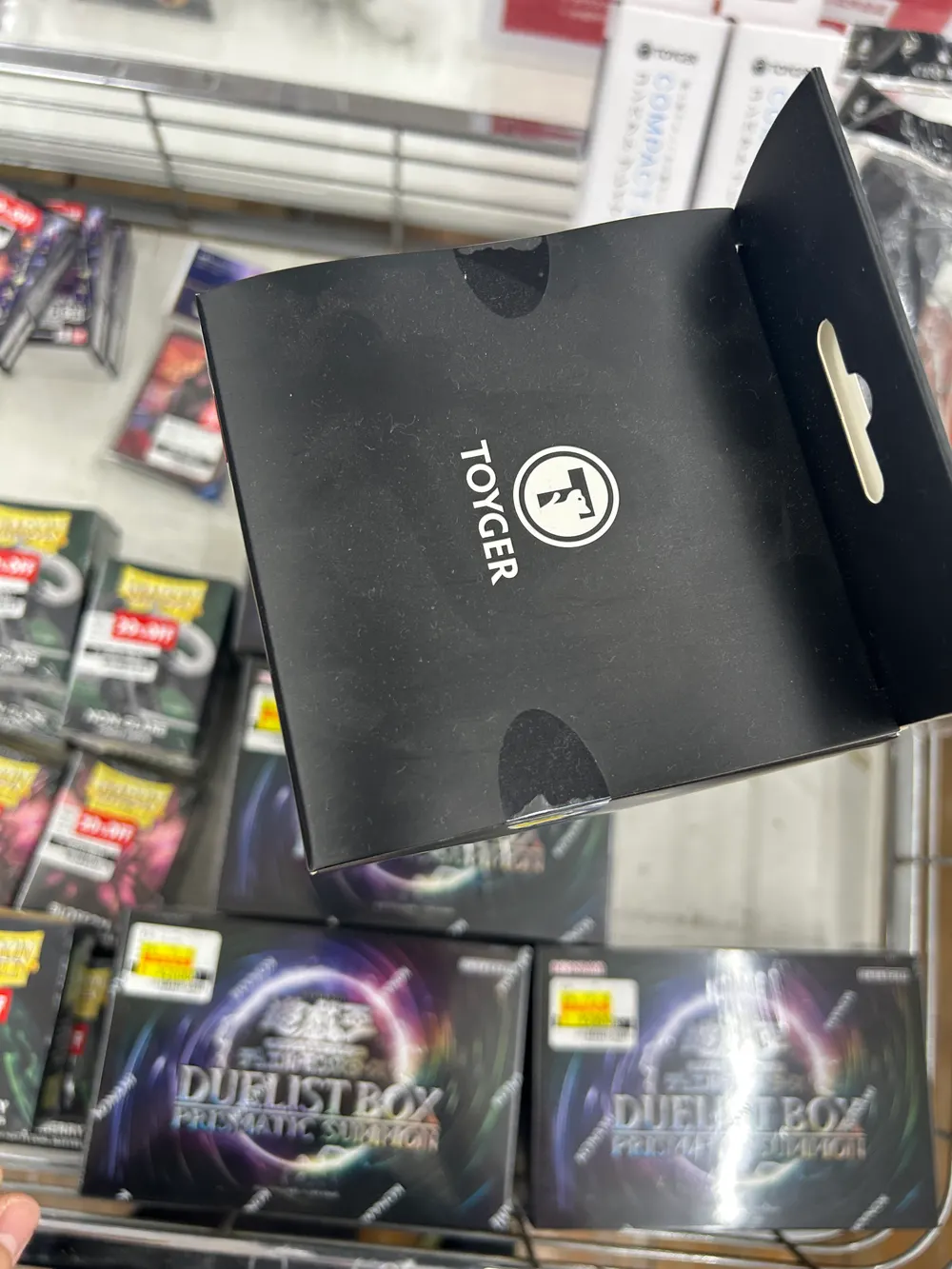 TOYGERのロゴが箔押しされた黒い箱を手に持っている写真。背景のワゴンには遊戯王のDUELIST BOXと50%OFFラベルの付いたスリーブが並んでいる。