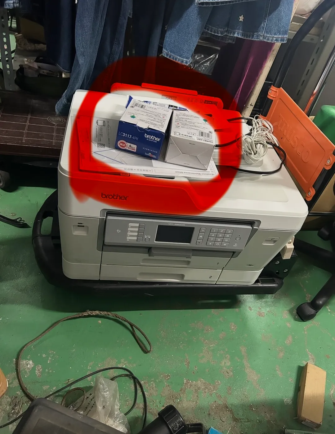 仕入れた白いブラザーの複合機プリンター MFC-J6983CDW を、倉庫の緑色の床の上で撮影した写真。本体の天面に、青と白のパッケージの「LC3117-4PK インクカートリッジ 4個入」の箱と、白い純正部品の箱、取扱説明書やメンテナンスシートなどの書類がまとめて載っており、その部分が赤いペンで大きく丸く囲まれている。奥には作業場の衣類ラックとオレンジ色のTRUSCO台車が写っている。