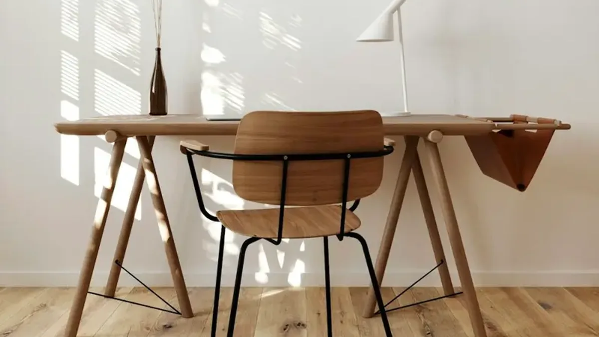 白い壁の部屋に置かれた木の机と椅子。窓からの自然光が床に伸びる、片付いた作業スペース。