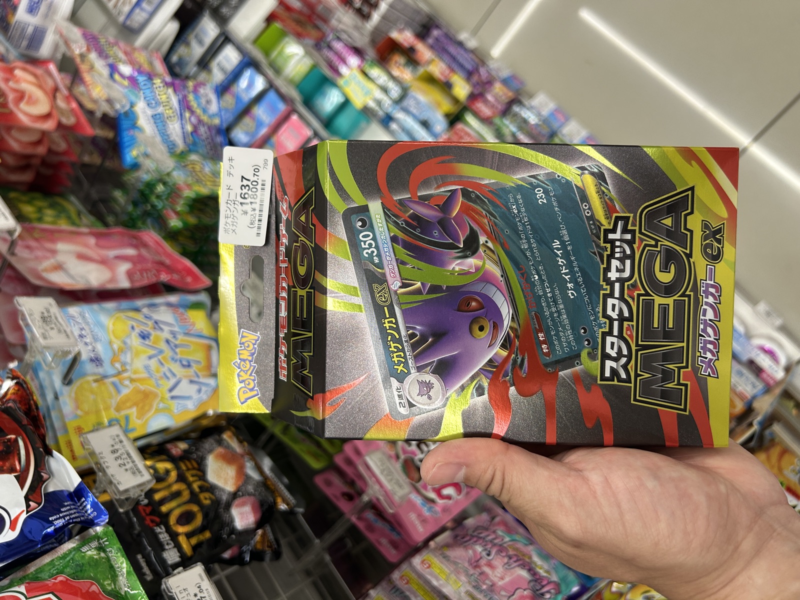 セブン-イレブン店内のお菓子コーナーで、ポケモンカードゲーム MEGAスターターセットex メガミュウツーexを手に取り、値札¥1,637（税抜）が見える様子