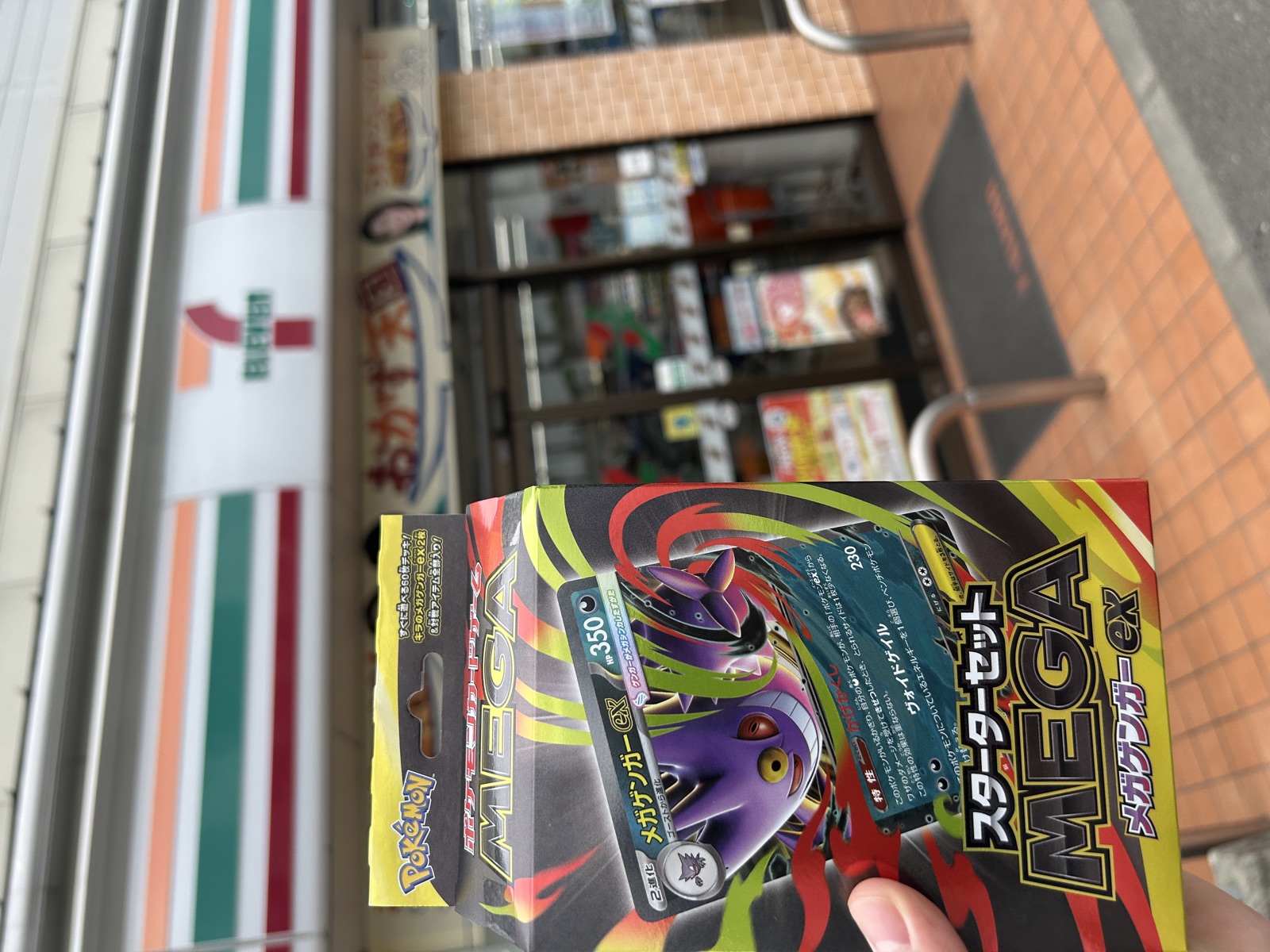 店外でセブン-イレブンの看板を背景に、2店舗で確保したポケカ MEGAスターターセットex 2個を手に持つ様子