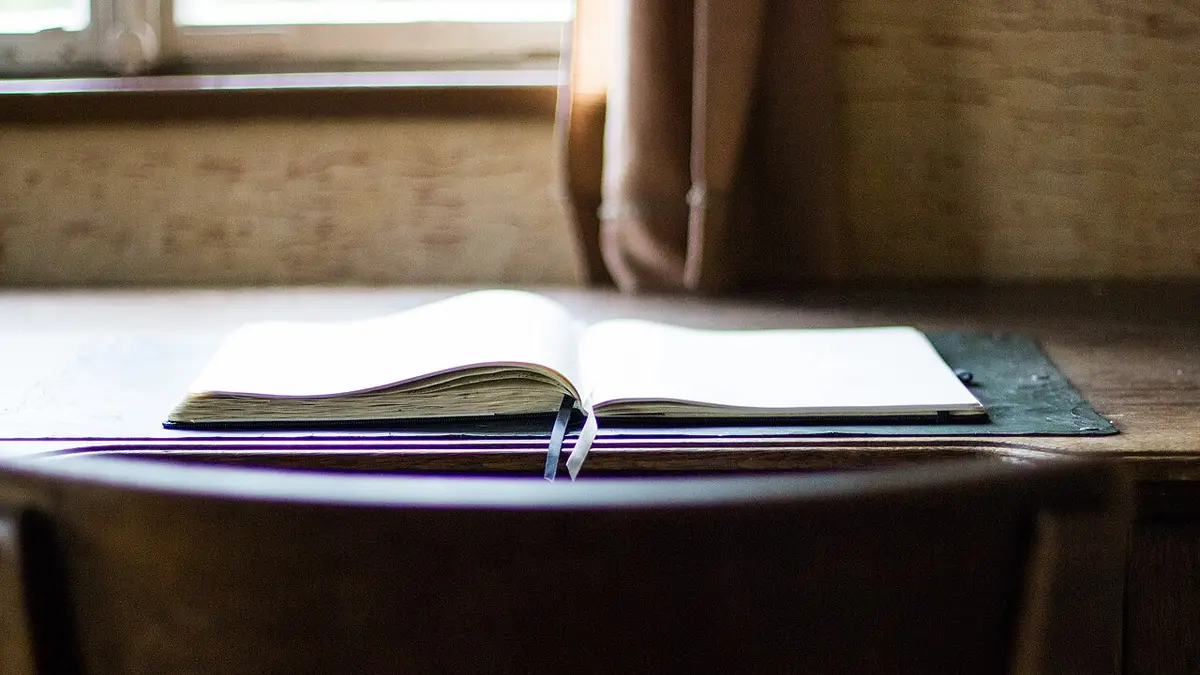 窓辺の木の机に開かれた本と、しおり代わりのペン。対話形式のやさしい入門書を読み進める場面