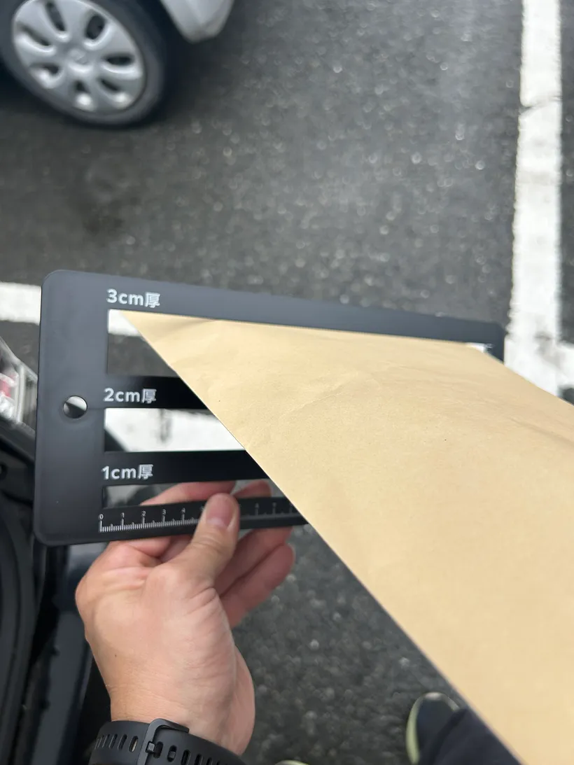 「3cm厚」「2cm厚」「1cm厚」の判定スリットが付いた黒い厚さ測定プレートと、クラフト封筒。駐車場で厚みを確認している。
