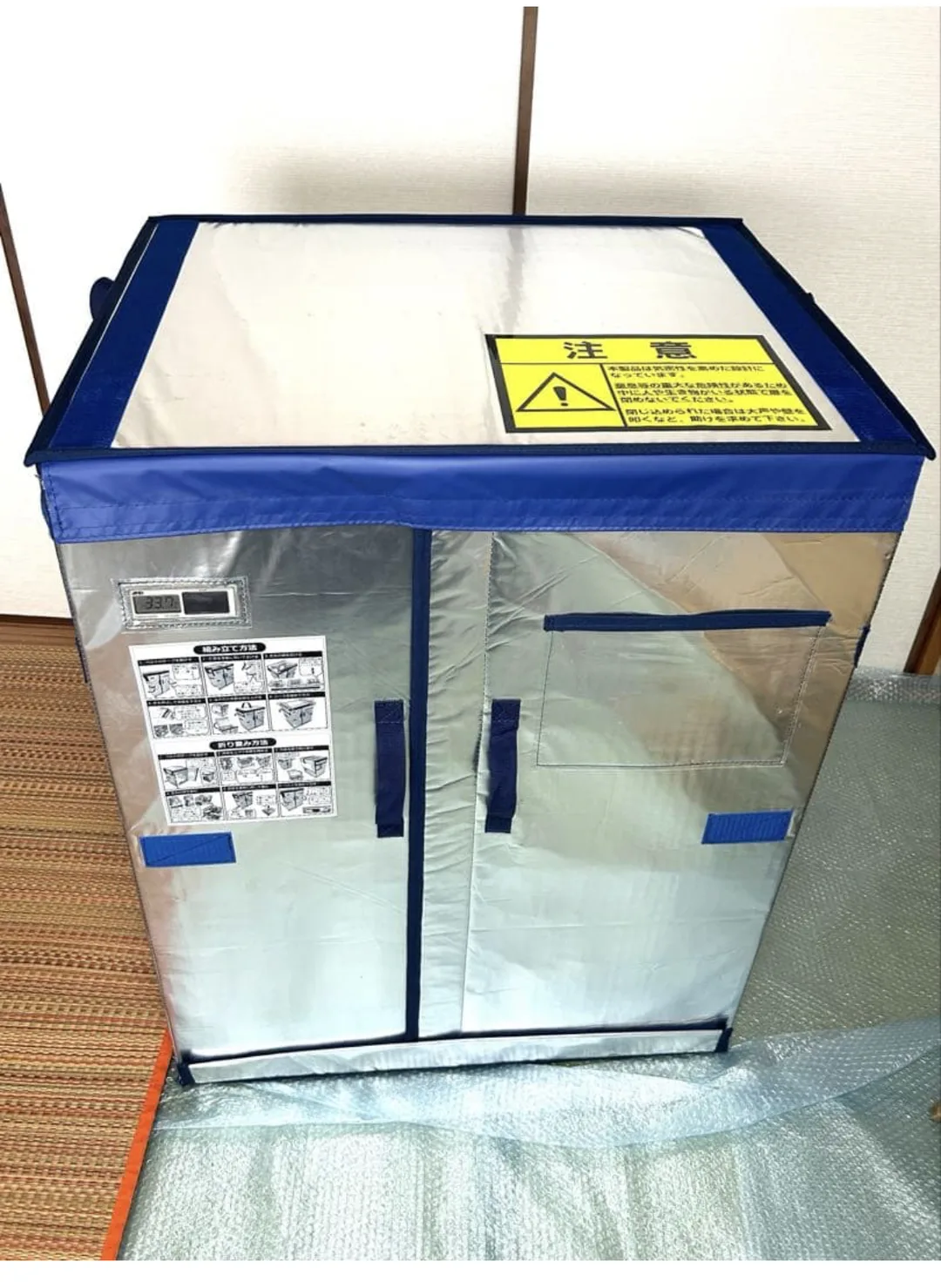 畳の部屋に置かれた、銀色の断熱シート製で紺色の縁取りが入った業務用の折りたたみ式保冷ボックス。天面に黄色い「注意」ラベル、前面にデジタル温度計と組み立て方法・折り畳み方法のイラスト説明シールが貼られ、観音開きの扉と面ファスナーのベルトが付いている。床にはプチプチが敷かれている。